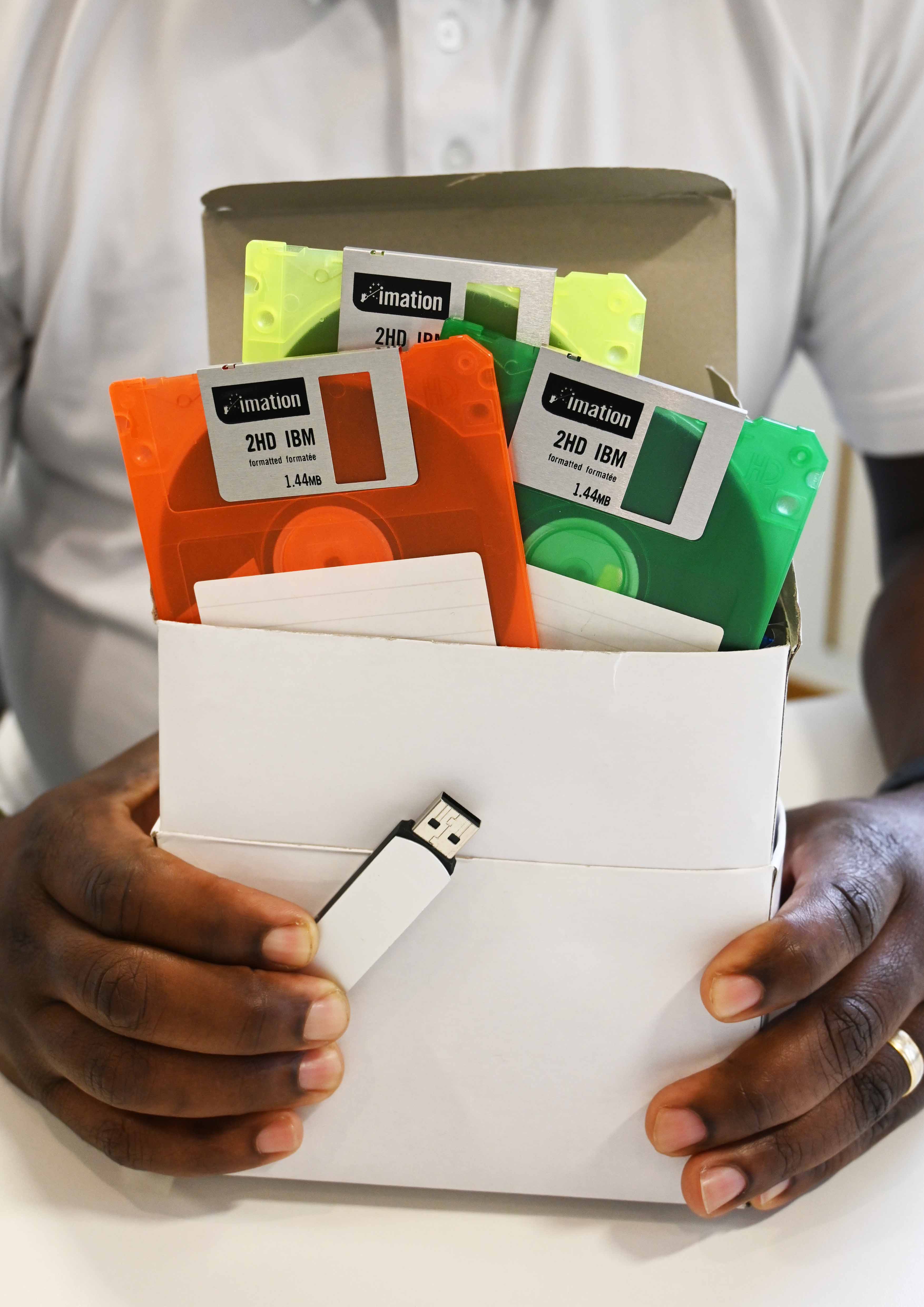 A man holds a white box in his hands. It's full to the brim with brightly coloured floppy discs - 726 to be exact. In front of the box in his other hand is a small USB drive. Both the box and the USB drive have exactly the same amount of memory. 