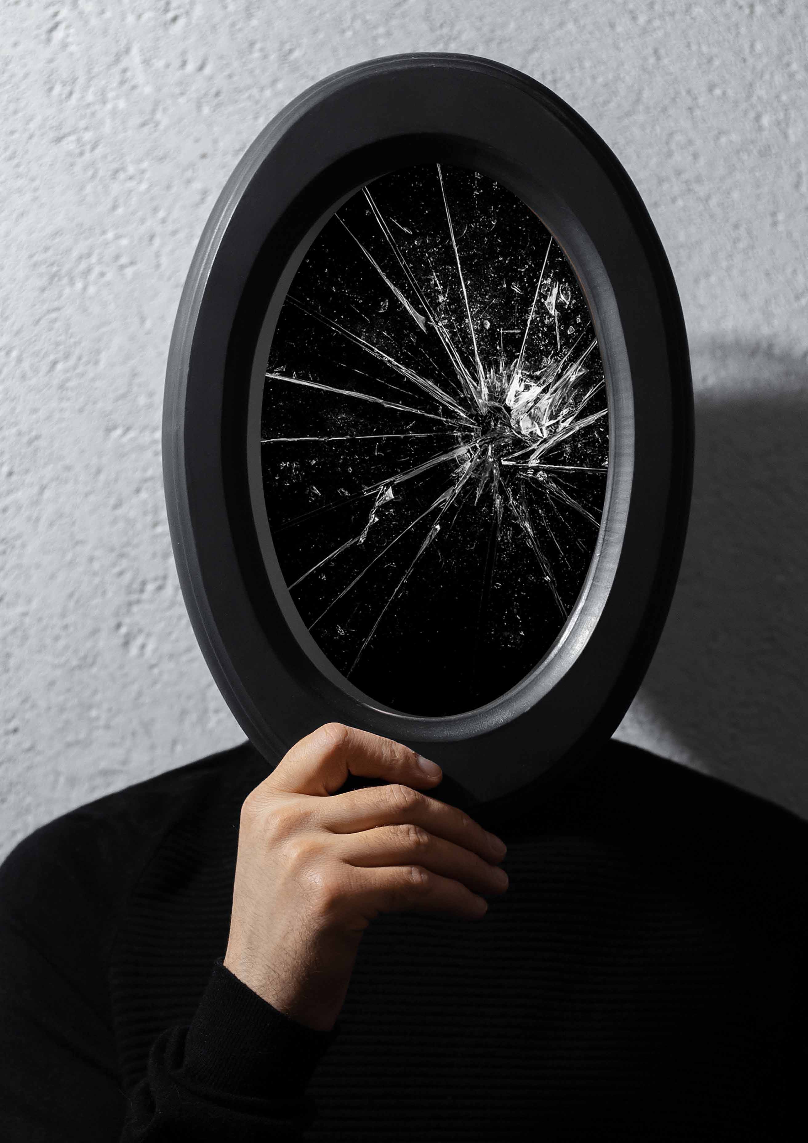 A person in a dark black outfit holds up a mirror to cover their face. The mirror is darkened and the glass is shattered.