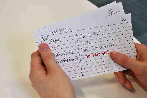 A person is holding a set of index cards. The index card lists an electrician, named Joe Smith. It has his name, age, whether he is a member of union and a recommendation on whether this person should be hired or not.