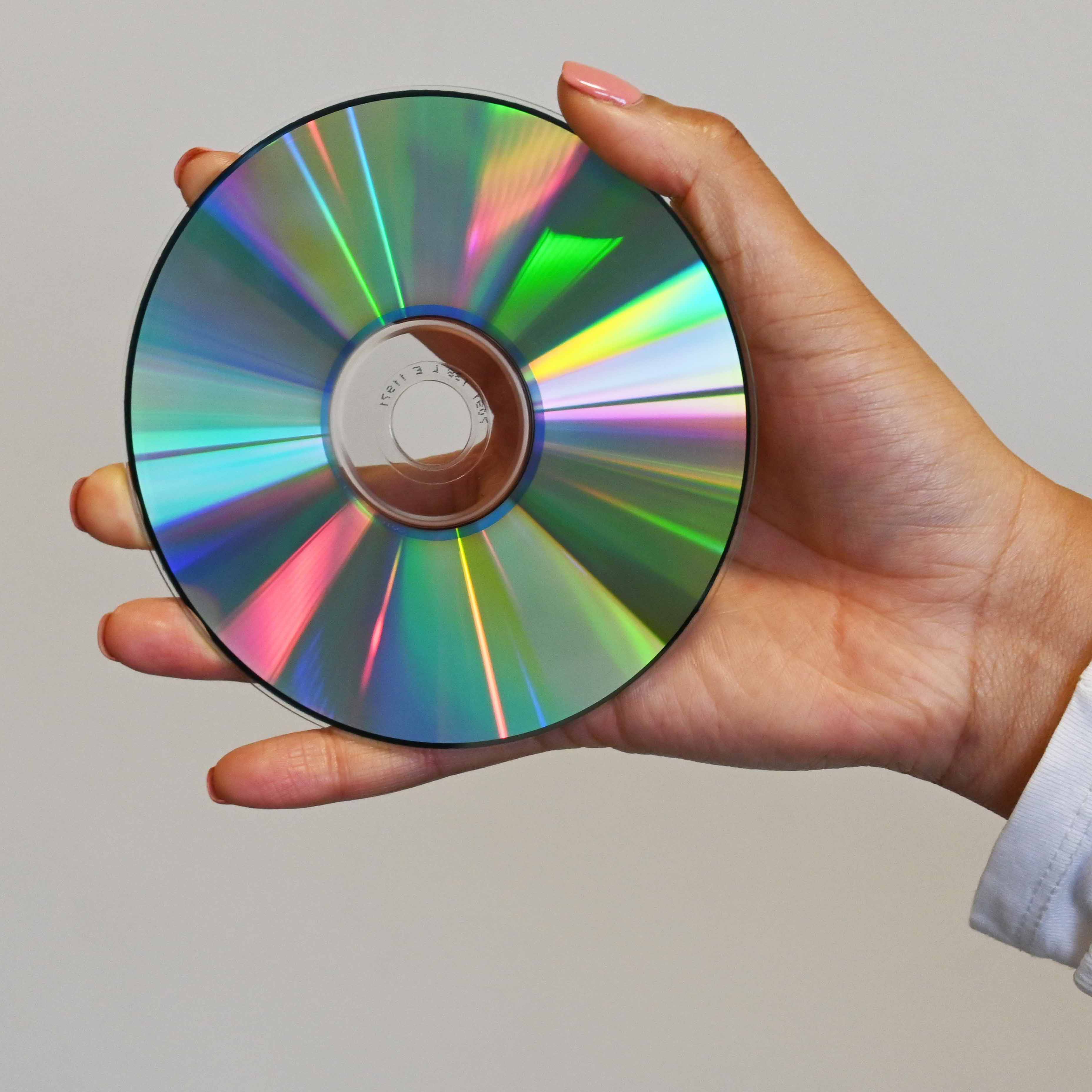 A woman holds a CD in the palm of her hand close up to the camera so you can see the rainbow holographic lights of the CD shine. 