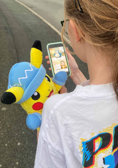 You can see over the over the shoulder of a child as she looks at her Pokémon Go game on her phone. She's carrying a Pikachu and wearing a Pokémon T-shirt.