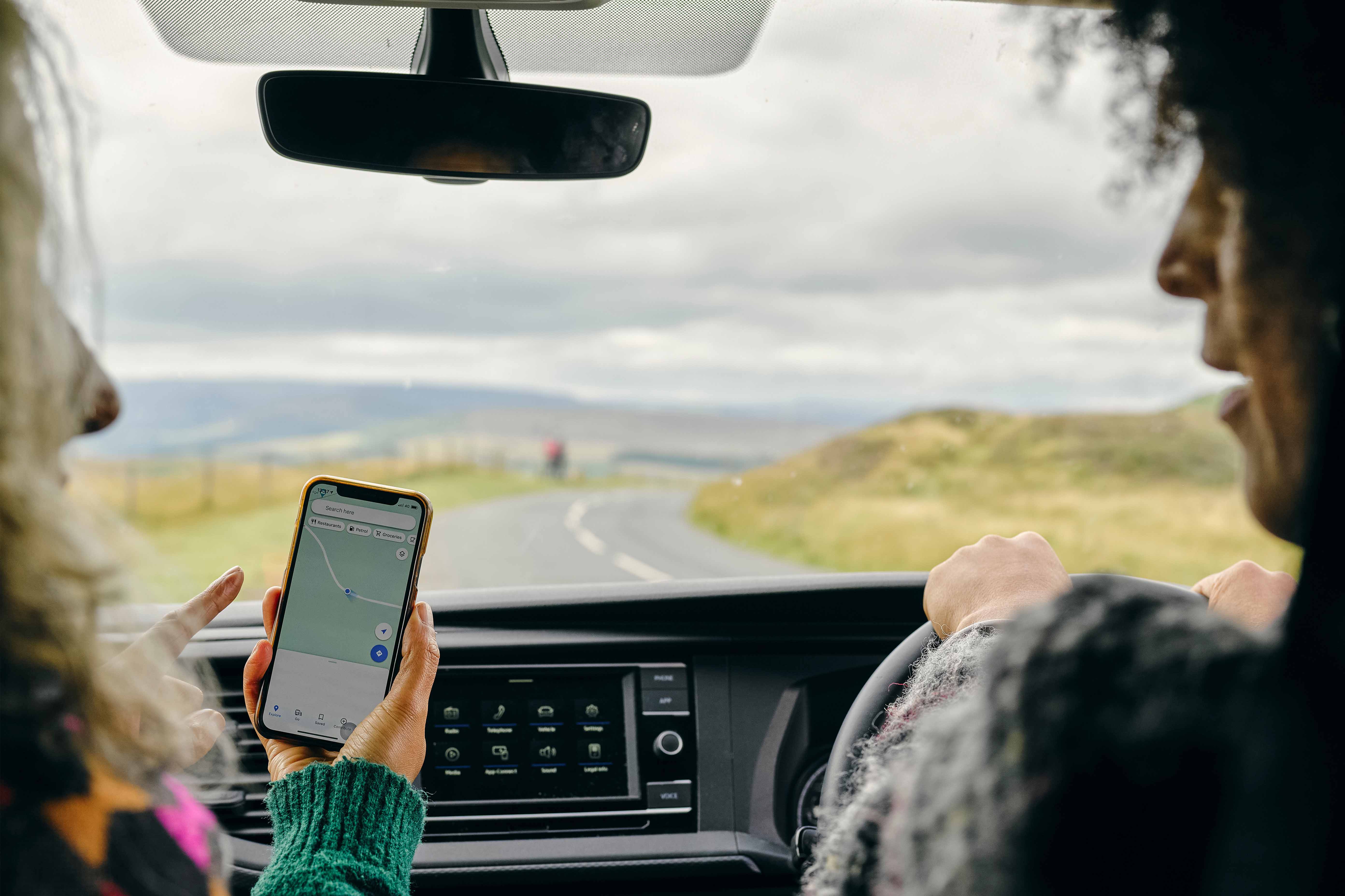 Two friends are chatting as they drive down a country road. The friend in the passenger seat points ahead as she looks down at Google Maps help her friend navigate the journey. 