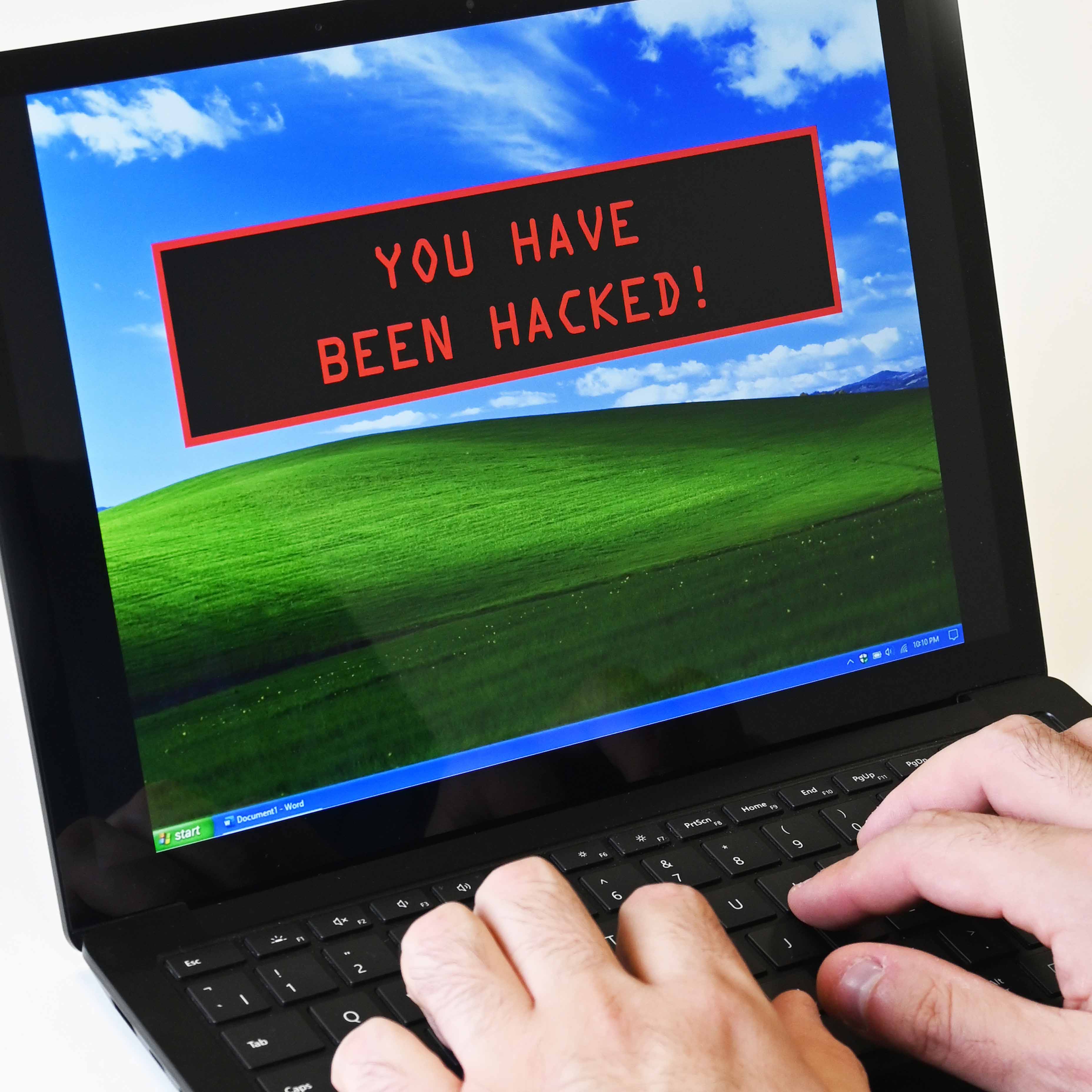 A man is typing at an old laptop. The desktop background on the screen is the classic windows background of rolling hills. However, over the top there’s a dark warning message. A black box has appeared and in a bright red robotic font the message “You have been hacked.” 