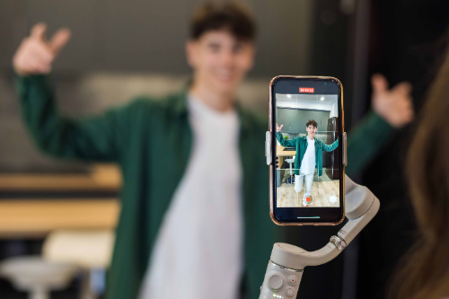 A young man has put his mobile phone in a stand and is recording himself doing a dance for social media.