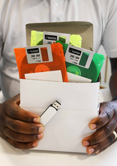 A man holds a white box in his hands. It's full to the brim with brightly coloured floppy discs - 726 to be exact. In front of the box in his other hand is a small USB drive. Both the box and the USB drive have exactly the same amount of memory.