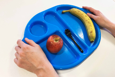 A person holds a blue school dinner tray. They've started to collect their dinner and have added a banana and an apple to the tray.