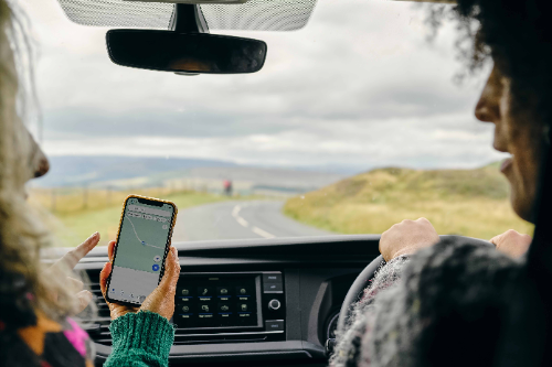 Two friends are chatting as they drive down a country road. The friend in the passenger seat points ahead as she looks down at Google Maps help her friend navigate the journey.