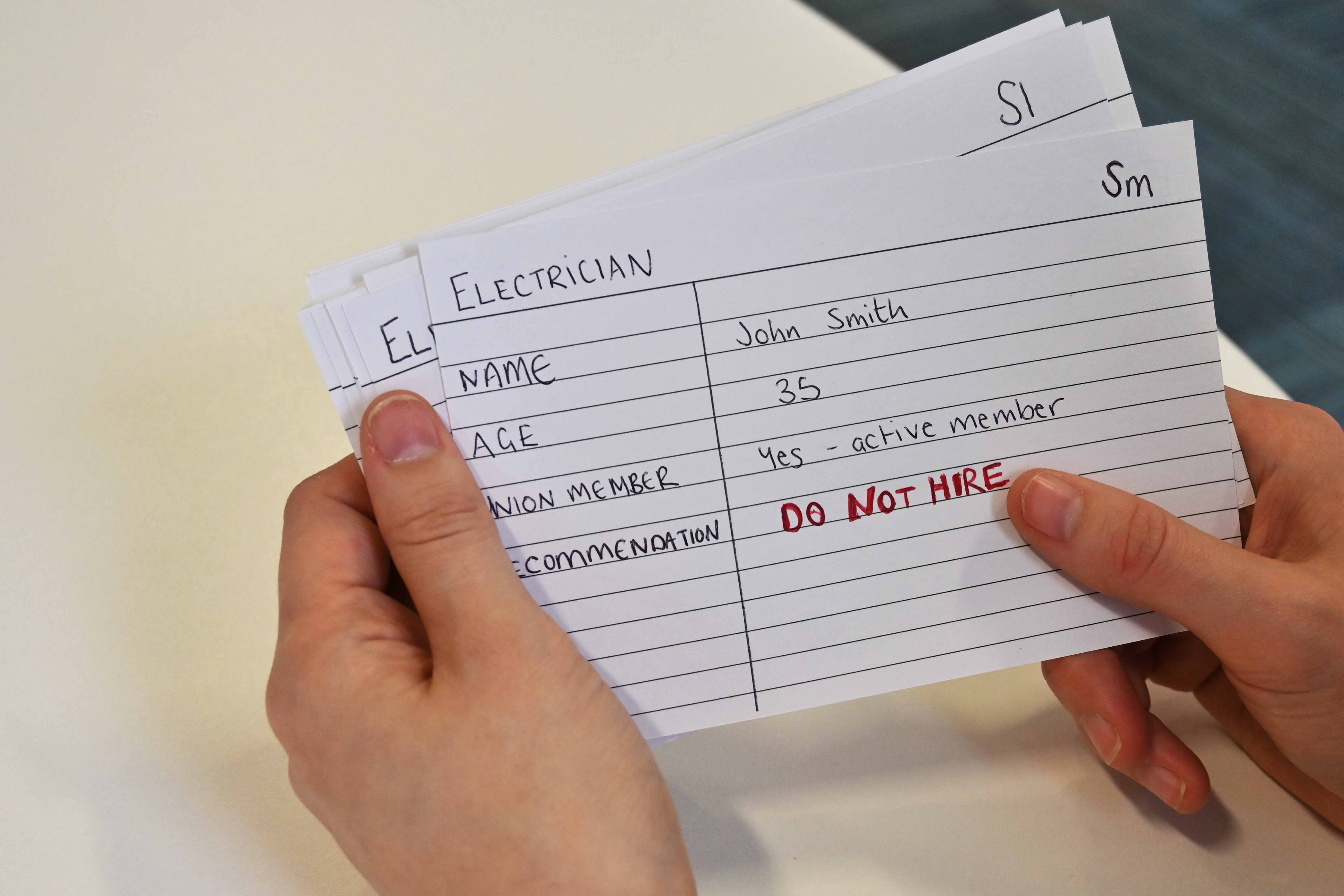 A person is holding a set of index cards. The index card lists an electrician, named Joe Smith. It has his name, age, whether he is a member of union and a recommendation on whether this person should be hired or not.