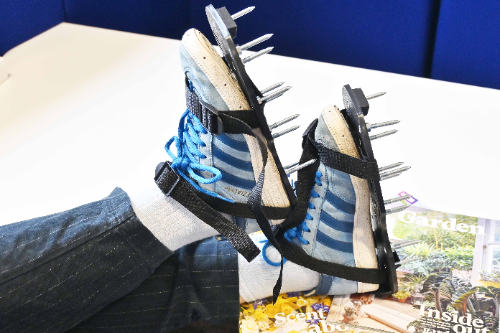 A woman has her feet up on a table. She’s leaning her feet on a pile of gardening magazines. She’s wearing blue Adidas trainers but added over the trainers are a pair of novelty grass aerator shoes. The shoes are attached to the bottom of the trainers and have large thick metal spikes out the bottom.