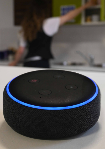 An out of focus image of a woman in the kitchen. She's leaning to get a mug from a cupboard. Closer to the camera you can see an Alexa smart speaker. It’s light is shining – showing that it’s listening and ready to answer the woman’s questions as she does other tasks.