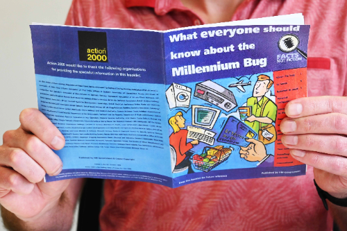 A man is holding and reading a small pamphlet about the Millennium Bug. The pamphlet has series of cartoons on the front representing shopping, flying, using technology like washing machines and computers, using credit cards and shopping. The title reads what everyone should know about the Millennium bug.