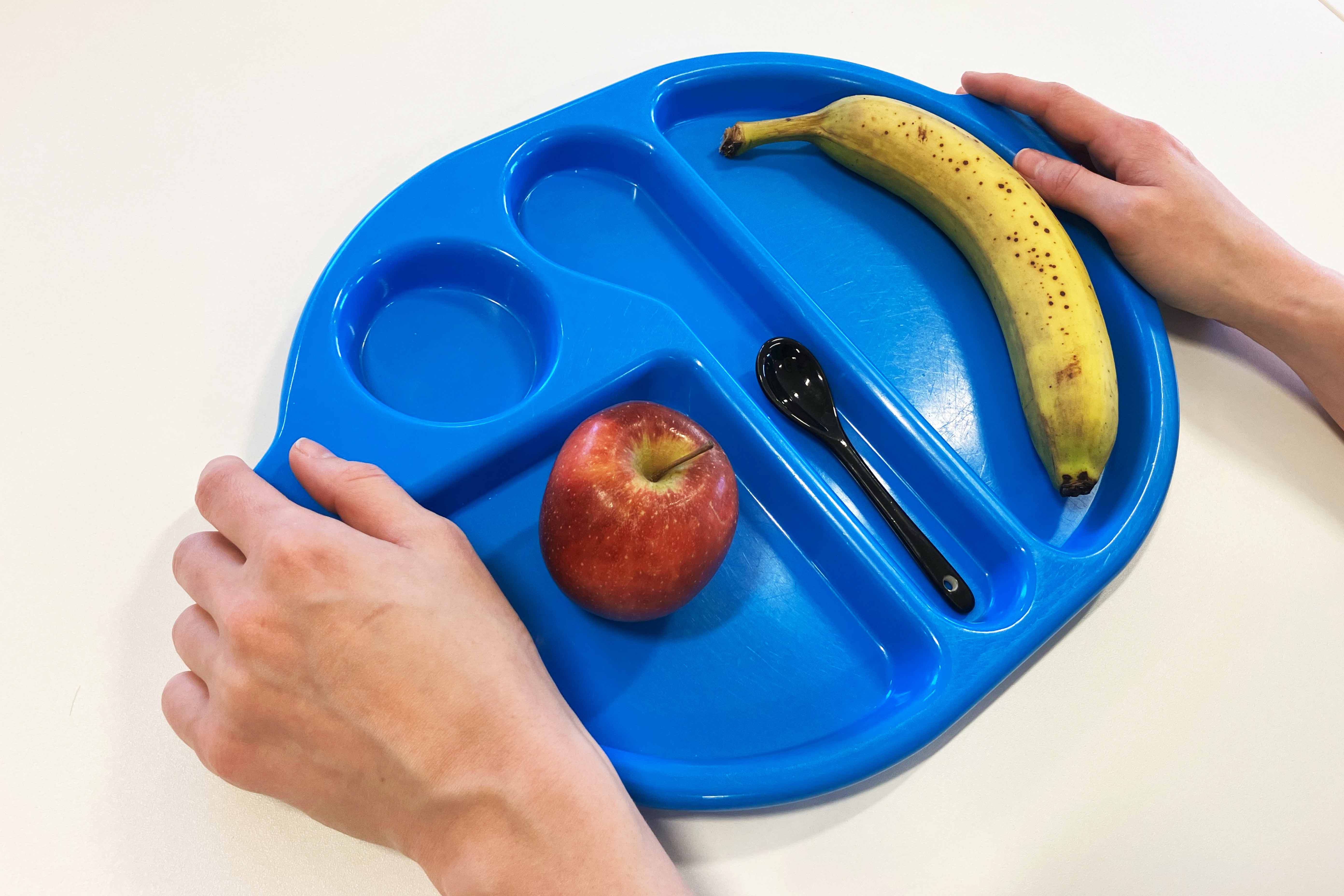 A person holds a blue school dinner tray. They've started to collect their dinner and have added a banana and an apple to the tray. 